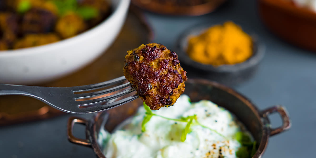 Indisk lammekjøttbolle på gaffel over en skål myntedressing med ferske urter på toppen. Indiske lammekjøttboller i hvit skål i bakgrunnen.