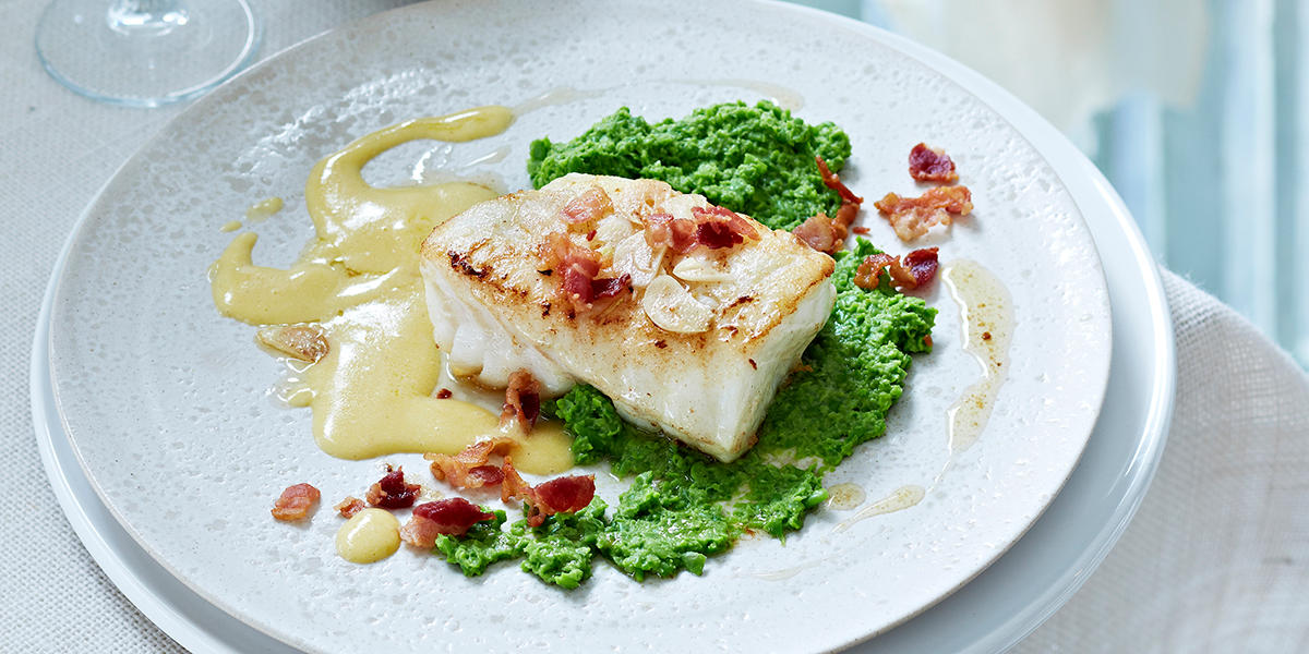 Pannestekt skrei med hollandaise og ertepuré servert på hvit middagstallerken på dekket bord, med sprøstekt bacon og skivet hvitløk over.