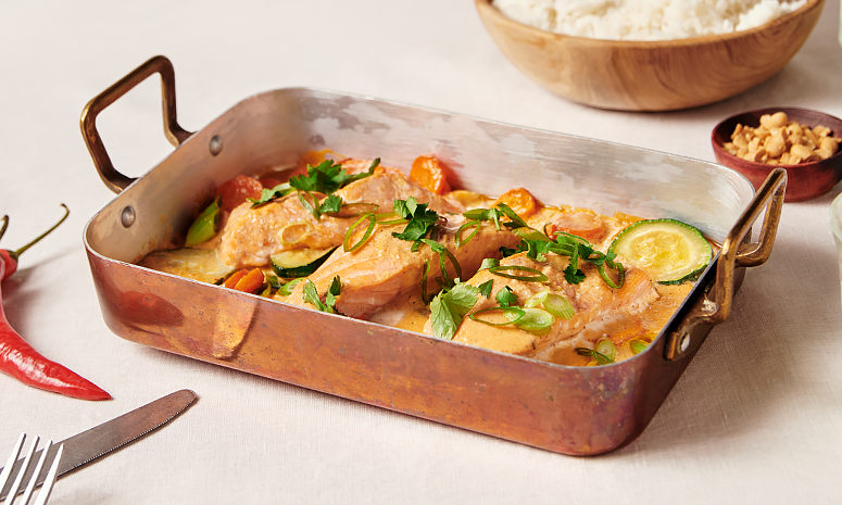Ovnsbakt laks i rød curry servert i ildfast form av messing, med grønsaker og ferske urter. I bakgrunnen står en trebolle med kokt ris, og en liten skål hakkede peanøtter.