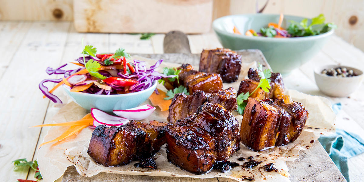 Asiatisk ribbe med soyaglaze servert i biter på trefjøl med bakepapir, og et dryss koriander over. I bakgrunnen er det flere blå og grønne skåler med syrlig fargerik coleslaw.