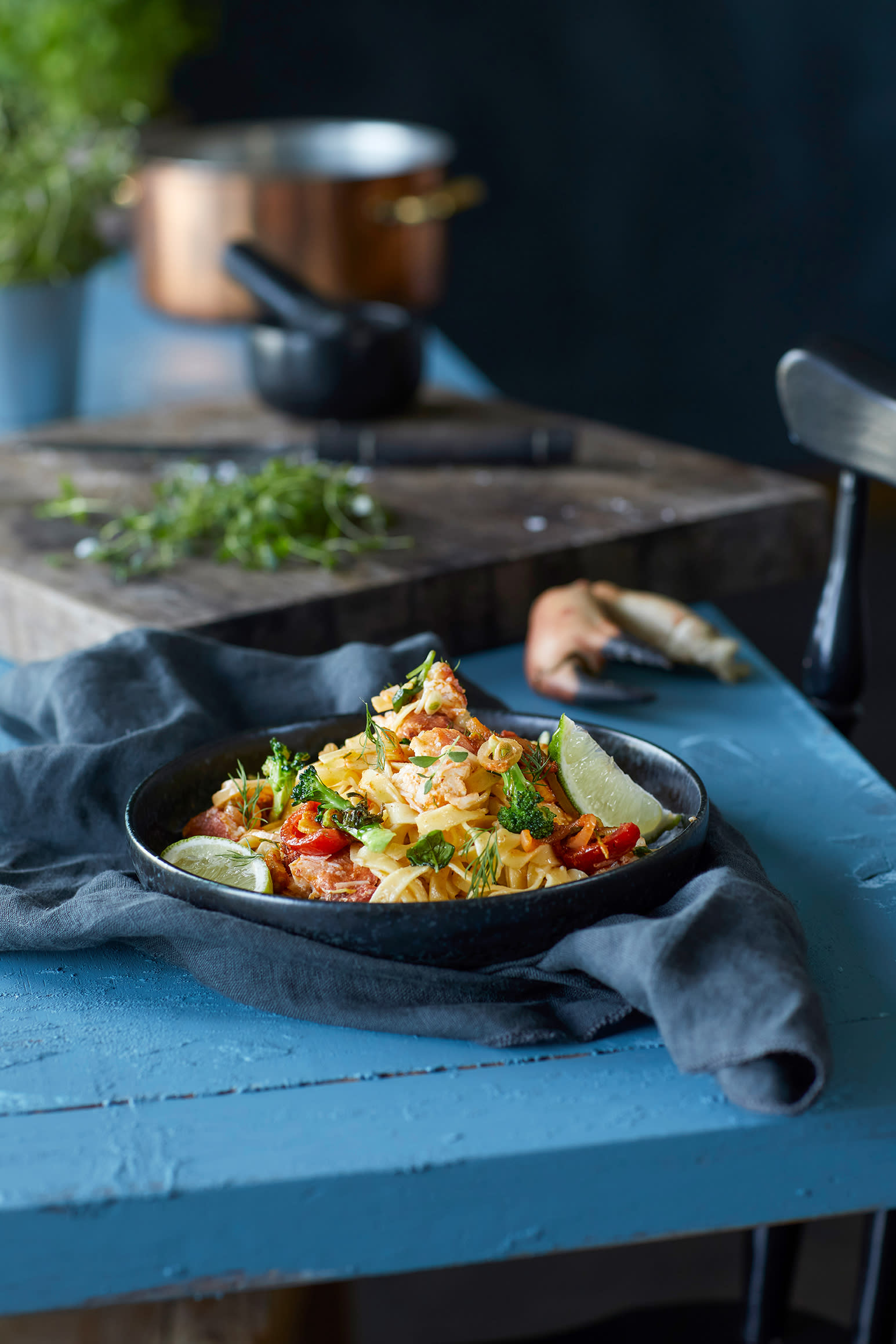 Tagliatelle med krabbeklør, tomat, brokkoli og creme fraiche, servert i svart skål med limebåter og frisk dill. Skålen står på rustikk blått kjøkkenbord med ferske urter, en gryte og en morter i bakgrunnen.