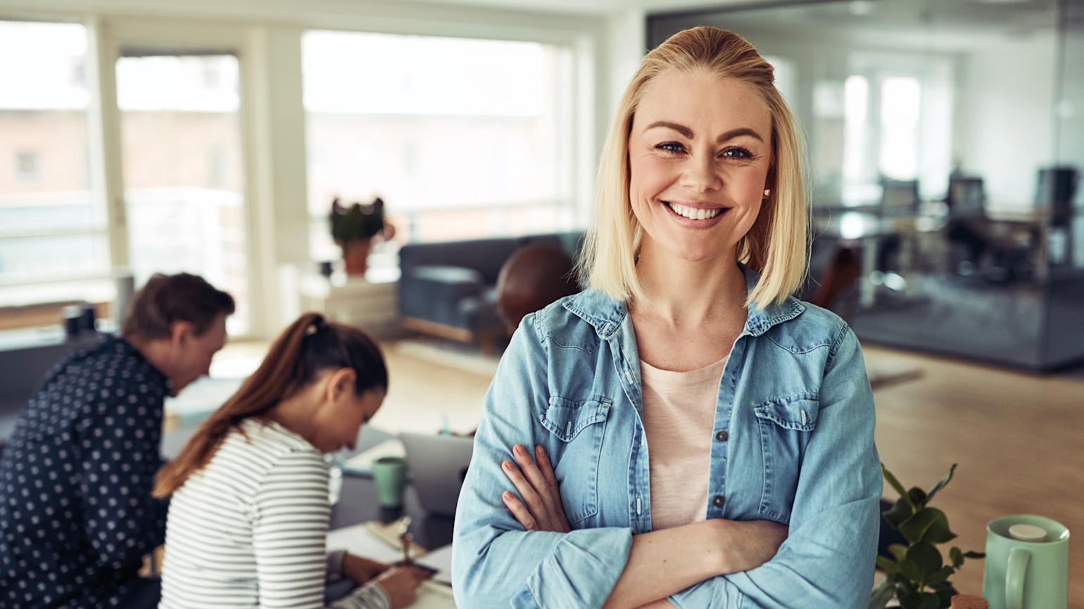 Blond kvinne smiler foran to som sitter og jobber i bakgrunnen