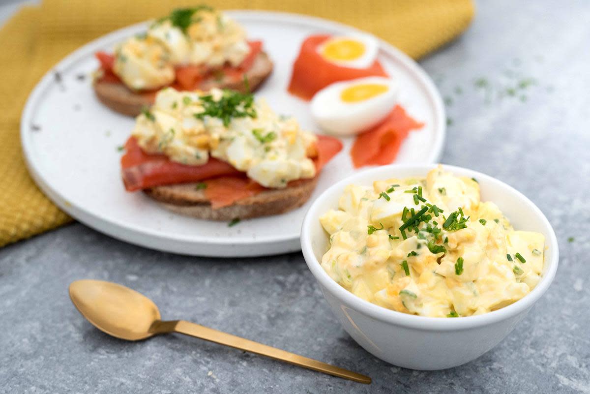 Eggesalat i hvit skål foran en tallerken med brødskiver med røkelaks, egg og eggesalat servert på grått bord med gult kjøkkenhåndkle ved siden av.