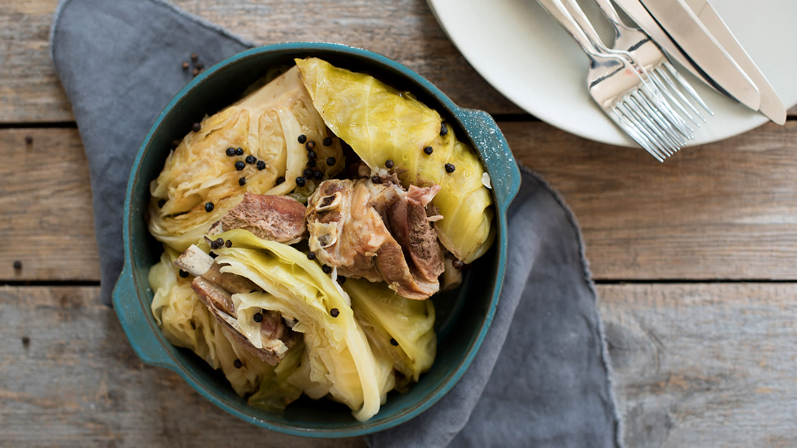 Tradisjonell fårikål med kål, lam og hel pepper servert i en turkis gryte på rustikk trebord. Mørkeblått kjøkkenhåndkle under, og bestikk ved siden av.