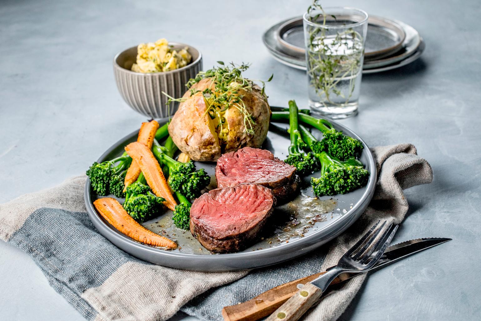 To skiver rosa biff servert på grå tallerken med bakt potet, brokkolini og gulrot på dekket middagsbord. Liten grå skål med kryddersmør i bakgrunnen.