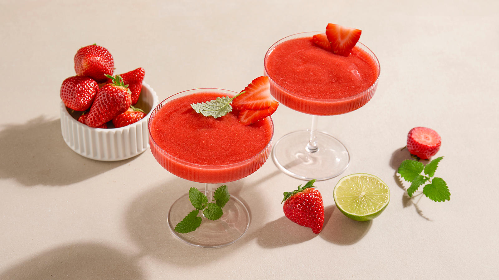 Strawberry daiquiri i to stettglass pyntet med friske jordbærskiver og grønne urter. Liten hvit skål med friske jordbær i bakgrunnen.