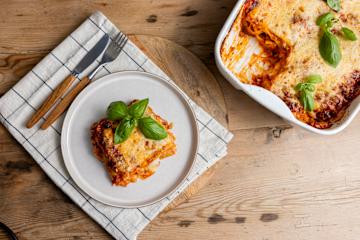 Enkel lasagne med høstens rotgrønnsaker laget av influenser Warunee, servert på hvit tallerken på rutete kjøkkenhåndkle. Tallerkenen står på rustikk trebord, med lasagneformen ved siden av, og bestikk i tre og metall på siden.