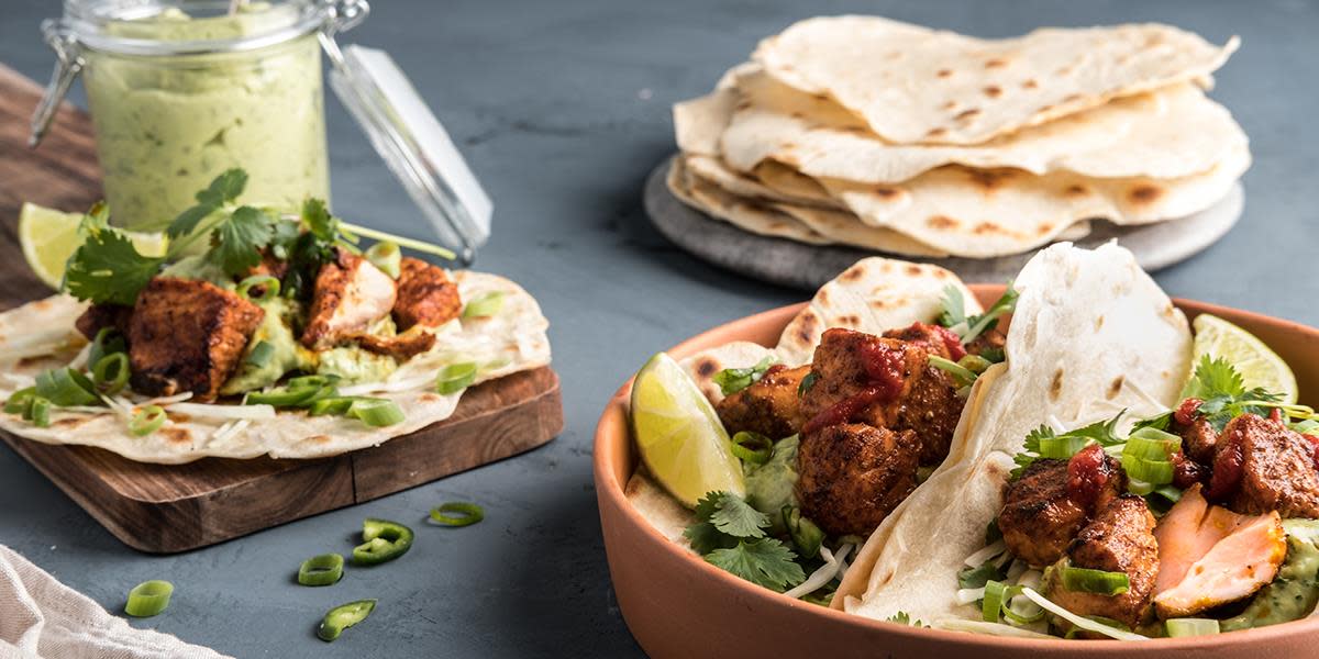 Laksetaco med avokadokrem og hjemmelaget tortilla servert i terracotta skål med frisk koriander og limebåter. Fat med hjemmelagede tortillas og krukke med avokadokrem i bakgrunnen.
