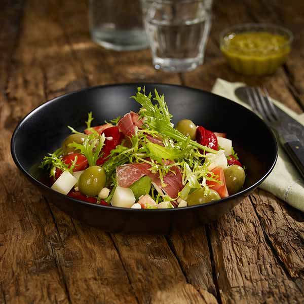 Spansk salat med Pata Negra, Manchego, oliven og tomater servert i svart skål på rustikk trebord.