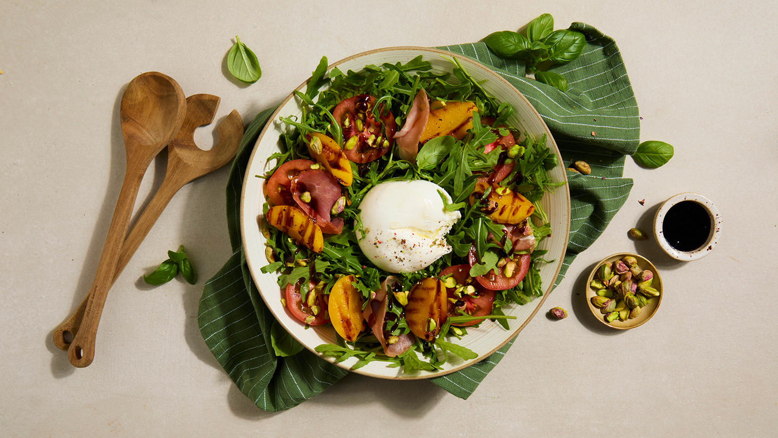 Sommersalat med grillet fersken, spekeskinke og burrata servert i lys bolle på grønt kjøkkenhåndkle med pistasjenøtter og salatbestikk i tre på siden.