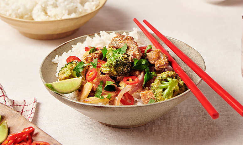 Svinekjøtt med rød curry og kokosmelk servert i en skål med røde spisepinner og en limebåt, i bakgrunnen er det en stor skål med ris