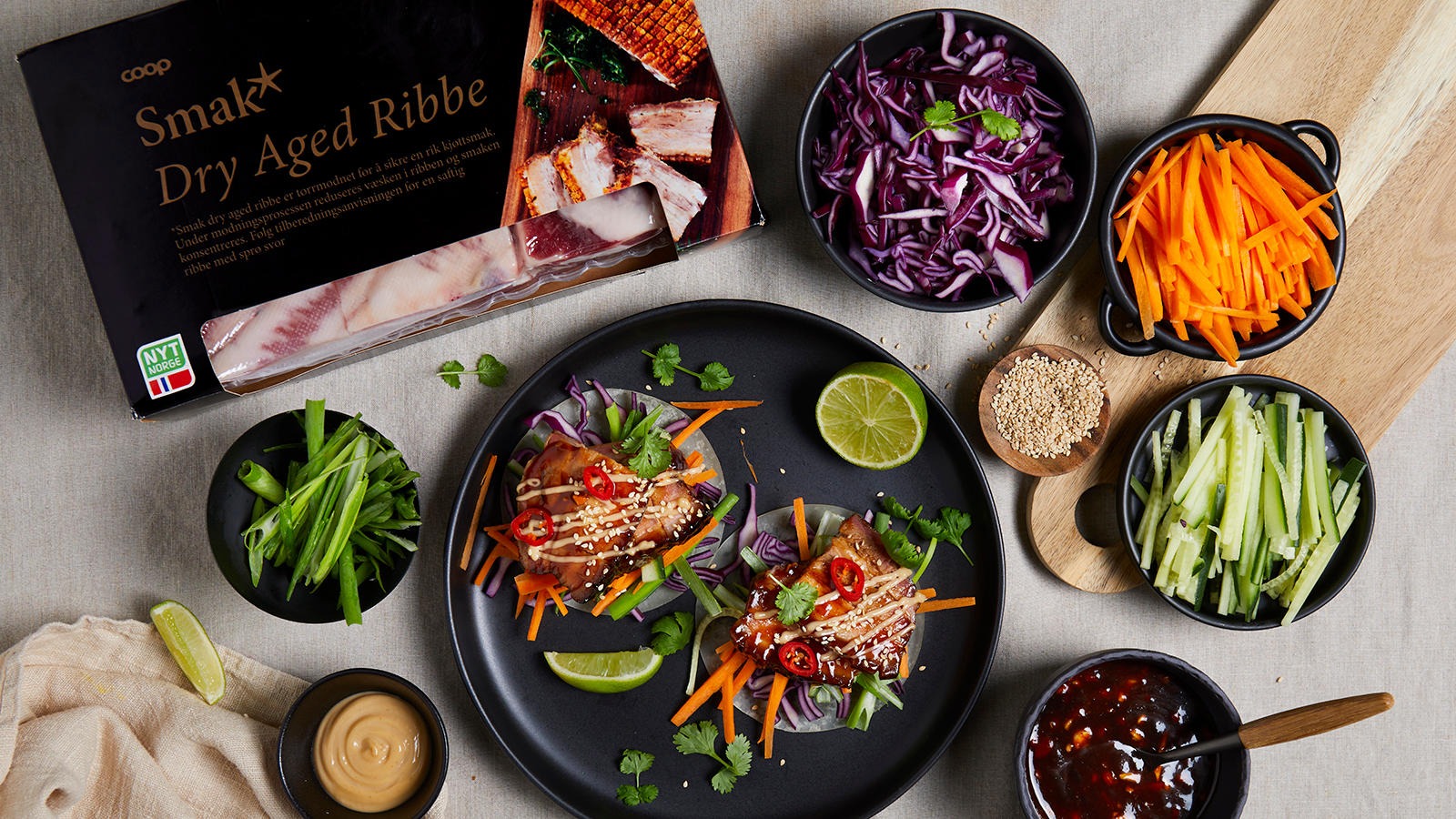 Asiatisk ribbe med tilbehør a la crispy duck servert i asiatiske pannekaker med finkuttede grønnsaker og hoisinmayo på svart tallerken. Små skåler med grønnsaker og sauser rundt.