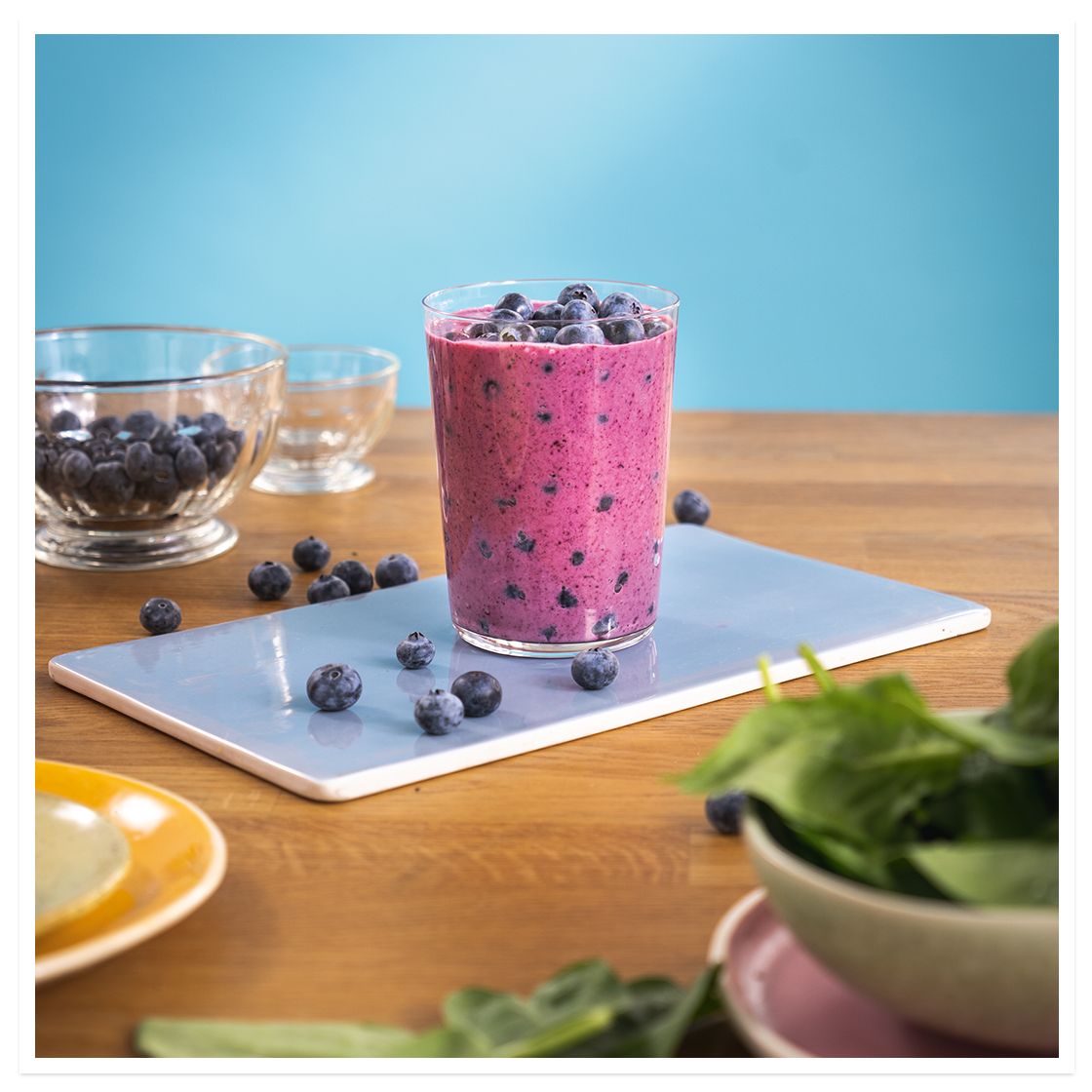 Himmelblå smoothie, lilla smoothie med blåbær og banan, i glass på blå fjøl på trebord. Ferske blåbær i bakgrunnen og på toppen av smoothien.