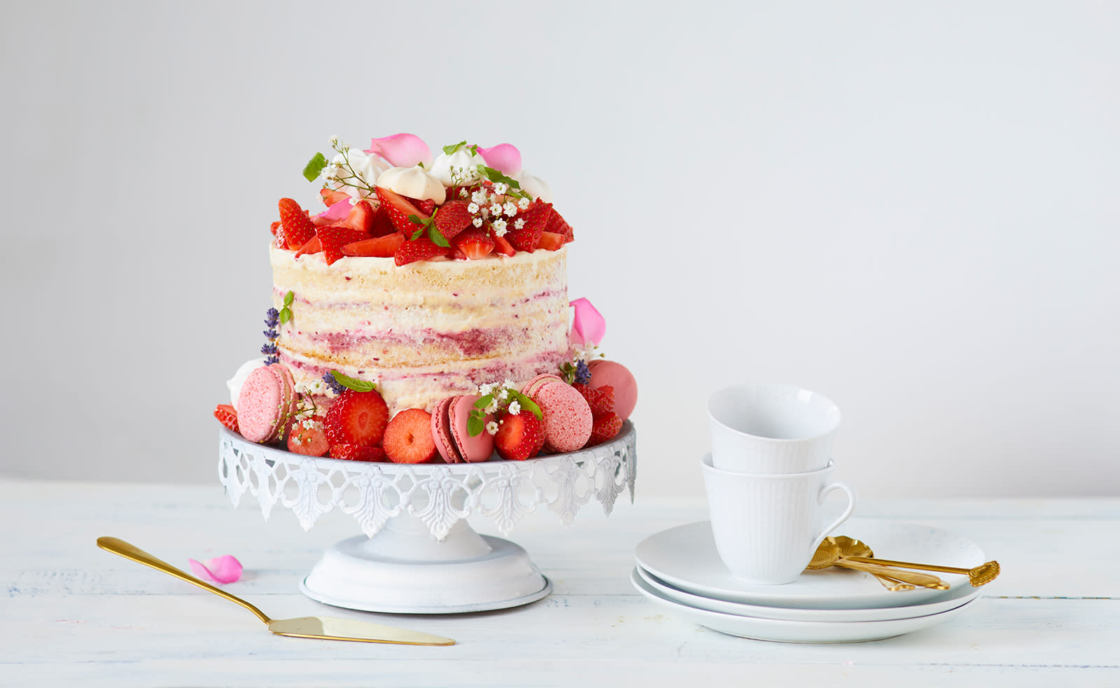 Bløtkake med pisket krem og friske jorbær, pyntet med jordbær, sommerblomster, rosa makroner og et tynt lag krem, servert på hvitt stettfat på hvitt trebord. Gullfarget kakespade, og hvitt kaffeservise på siden.