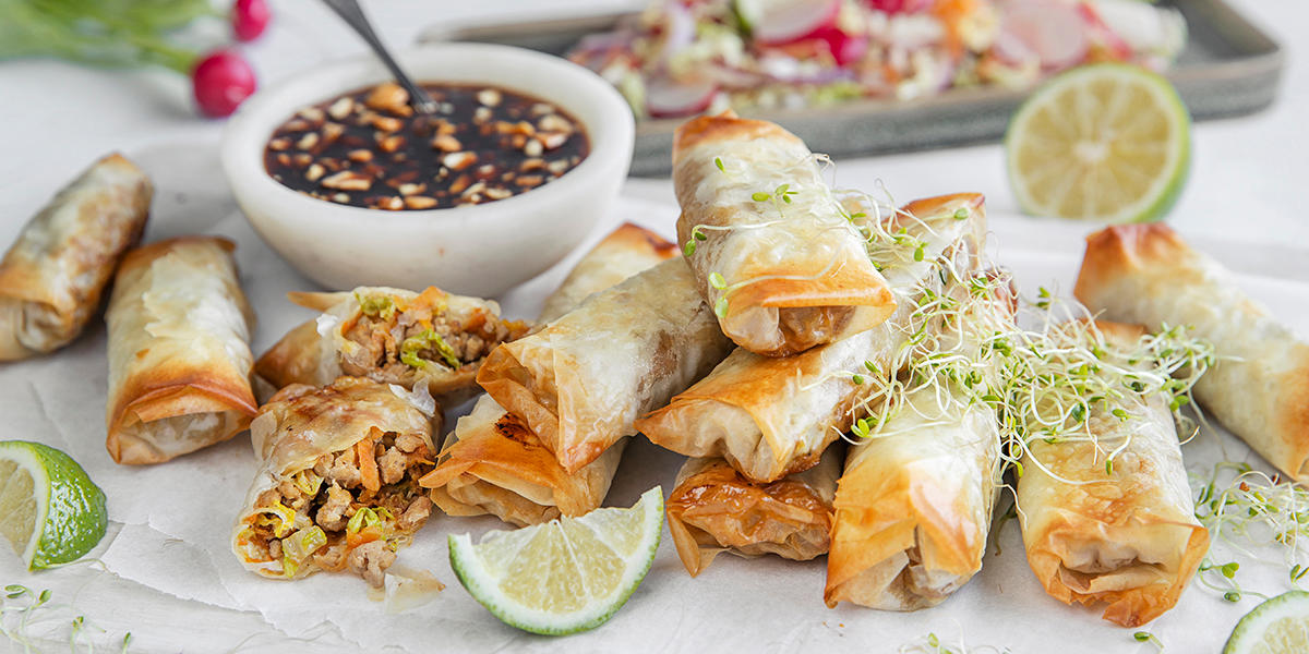 Ovnsbakte vårruller servert i stabler på hvitt bakepapir med bønnespirer på toppen og limebåter ved siden av. Soyadip i liten skål ved siden av, og brett med asiatisk kålsalat i bakgrunnen.