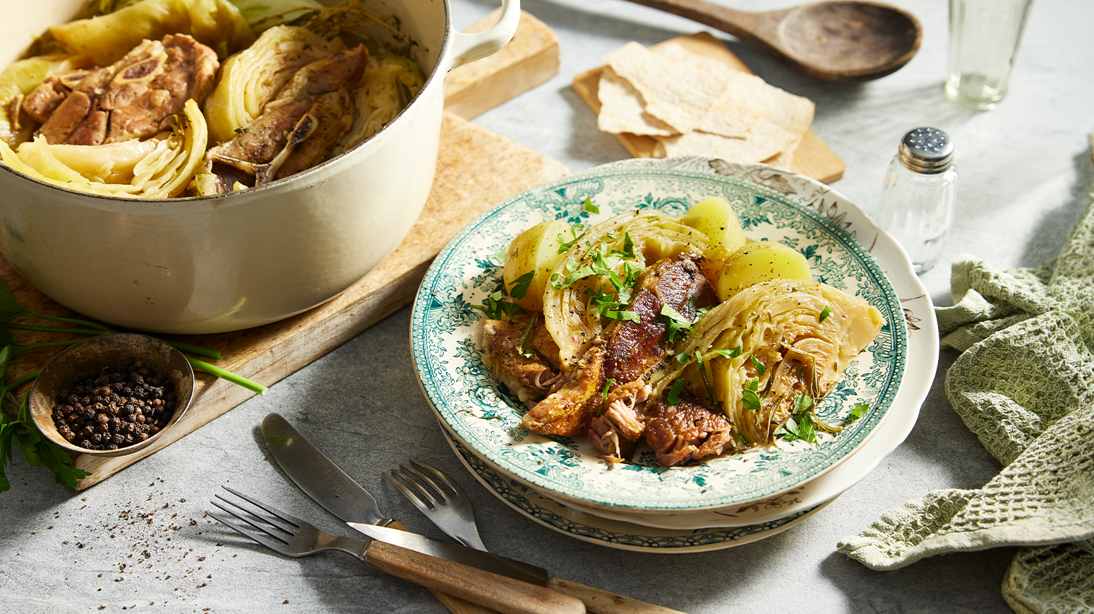 Fårikål med båter av kål og finhakket frisk bladpersille servert i grønnmønstret keramikkasjett på dekket middagsbord. Gryte med fårikål og liten fjøl med flatbrød i bakgrunn.