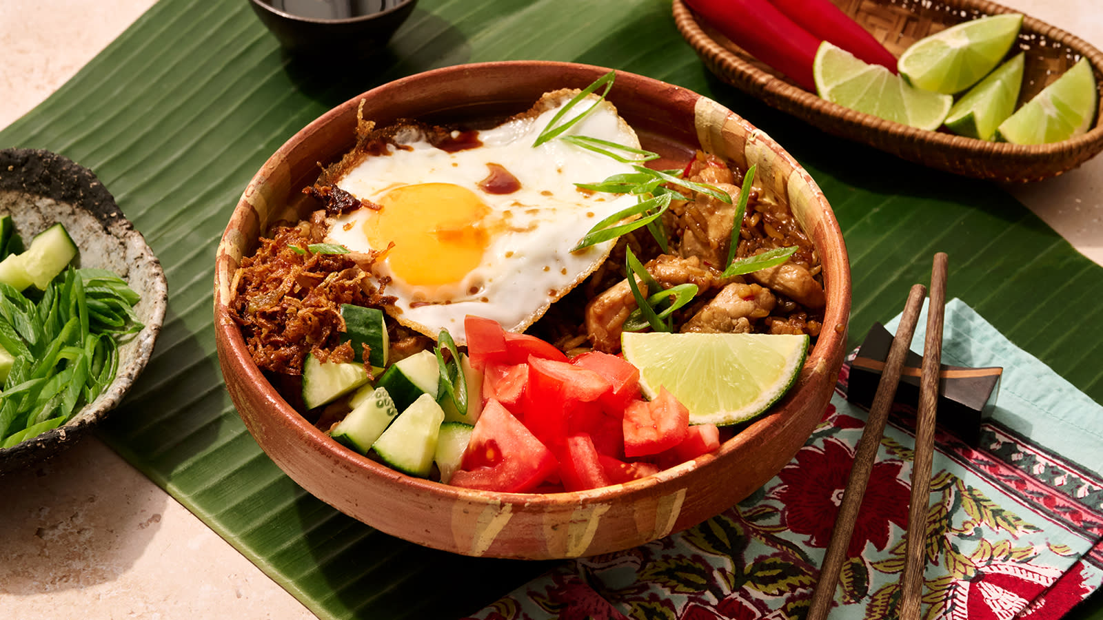 Nasi goreng - indonesisk stekt ris med kylling, servert med tomater og agurk i terninger, limebåt, sprøstekt løk og vårløker i strimler i en keramikkskål på bananblad.