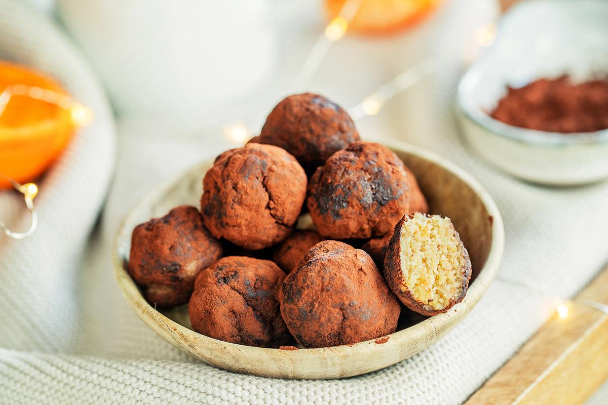Kakaorullede mandelkuler med dadler og klementin, servert i lys skål på lys stoffbakgrunn. Klementiner og julelys i bakgrunnen.