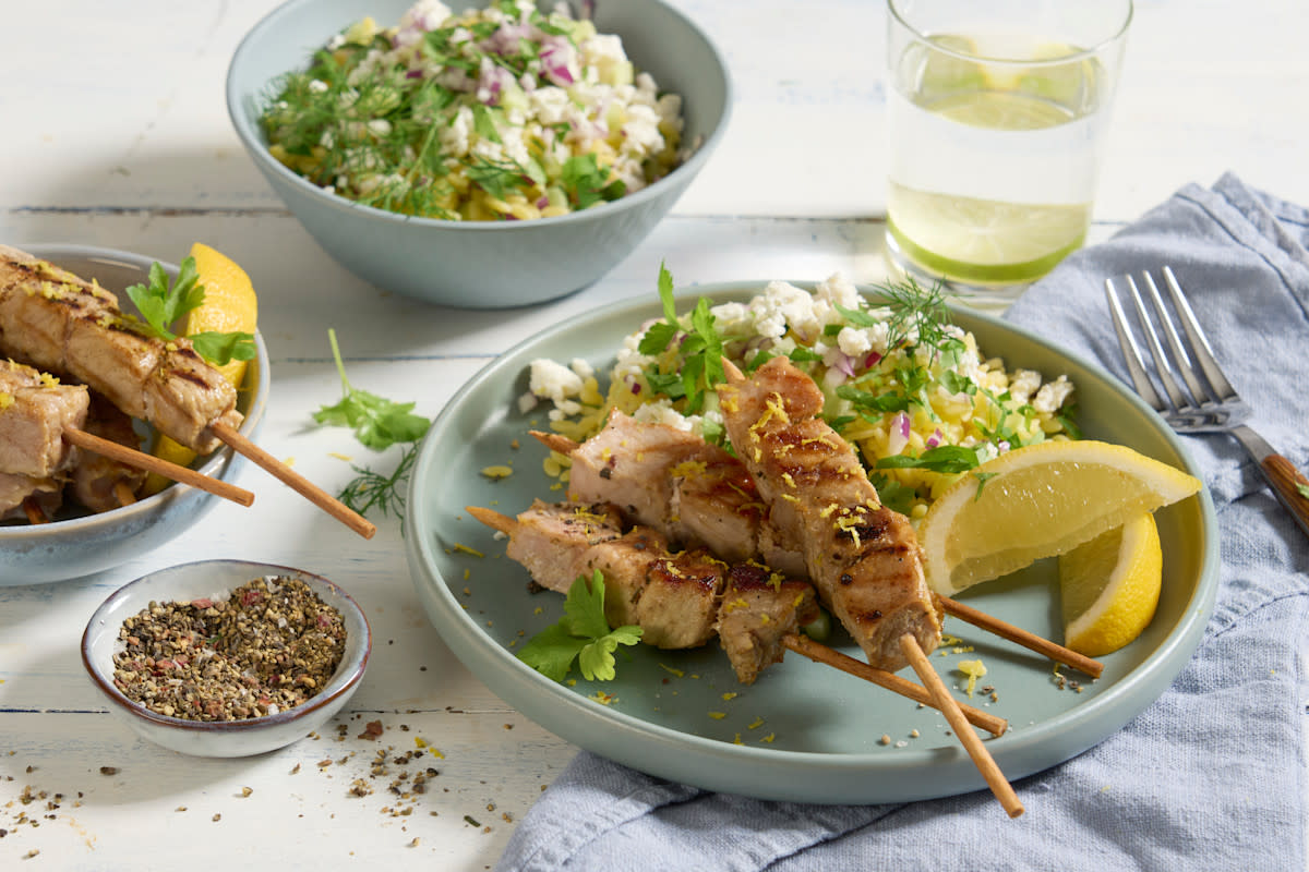 Spicy lemon grillspyd med gresk orzosalat servert på lysegrønn tallerken med sitronbåter og friske grønne urter. Lysegrønn skål med orzosalat, og vannglass med limeskiver i bakgrunnen.