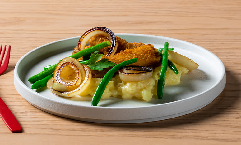 Fiskepinner med grov potetmos, aspargesbønner og stekt løk servert på en hvit tallerken med en rød gaffel og kvin ved siden av