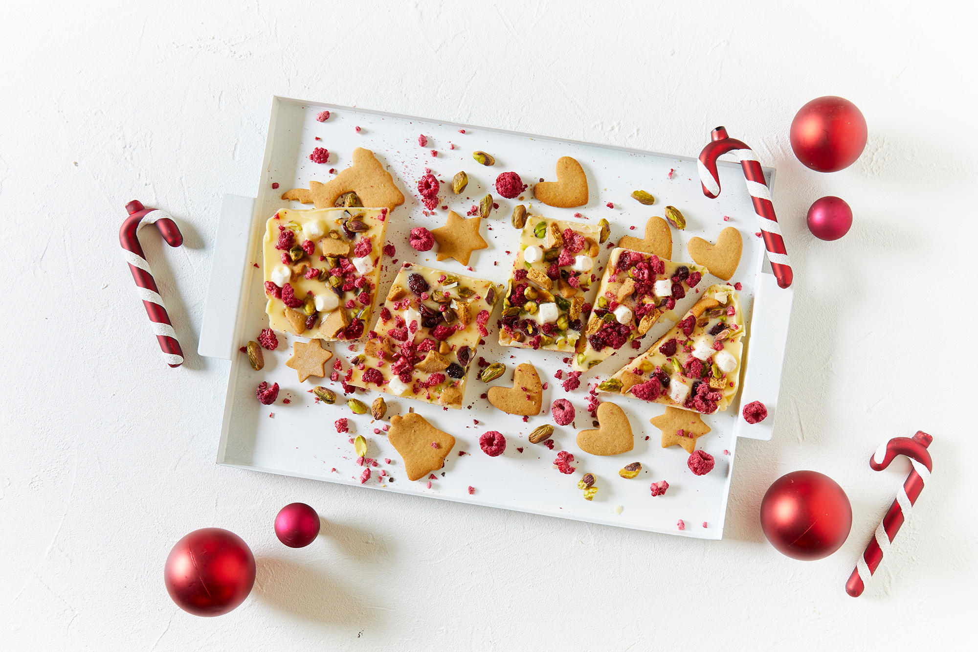 Hvit rocky road med bær og pepperkaker, delt i biter og servert på hvit firkantet tallerken med gylne pepperkaker og rød julepynt rundt.