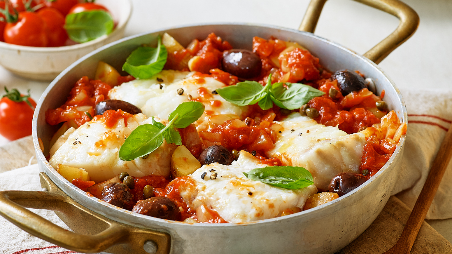  Et nærbilde av en torskegryte med tomat, oliven, kapers og fersk basilikum i en vid, kobberfarget gryte.