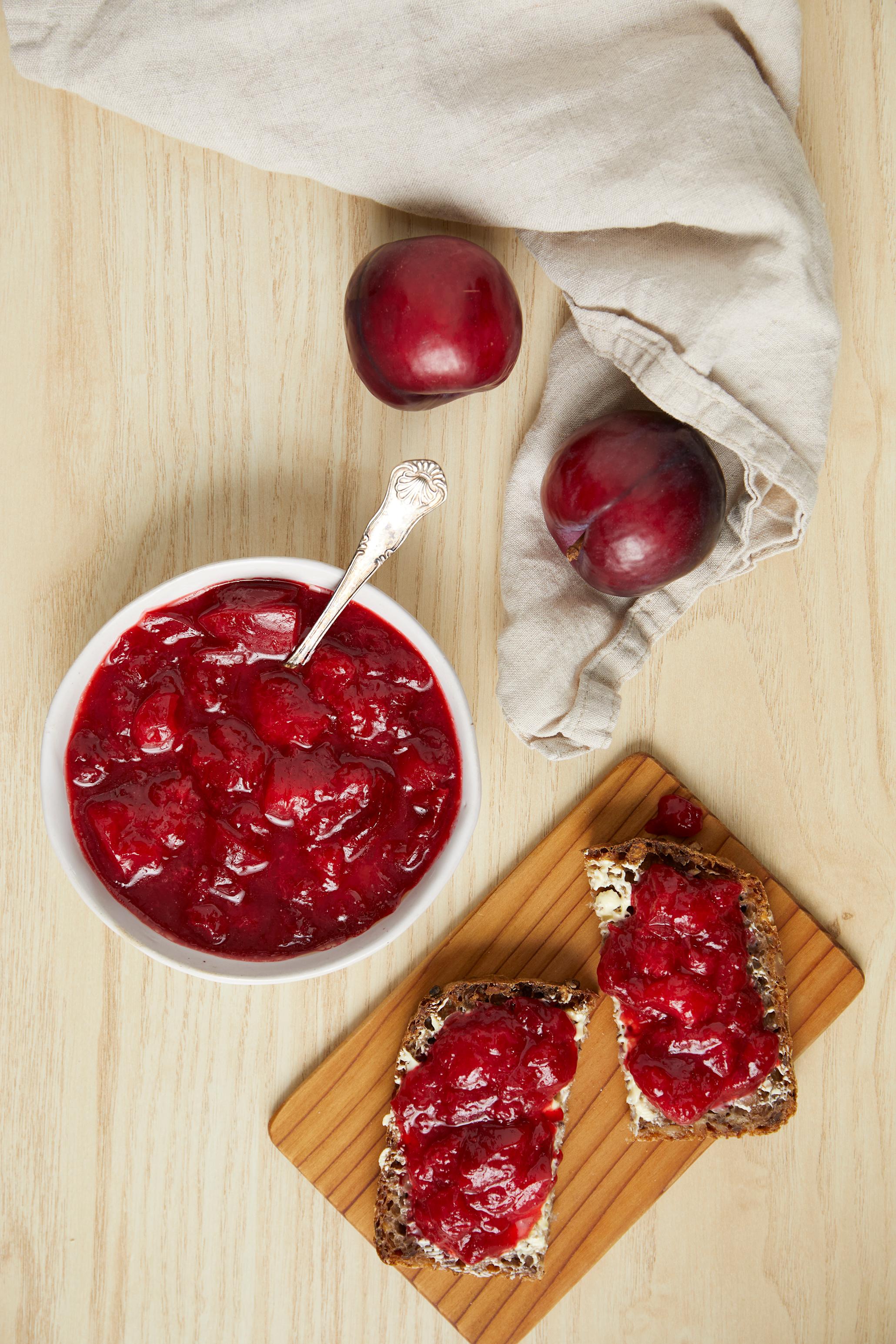 Plommesyltetøy med kanel og sitron, servert i hvit skål på lyst trebord, ved siden av trefjøl med brødskiver med smør og plommesyltetøy. Linduk med ferske plommer ved siden av.