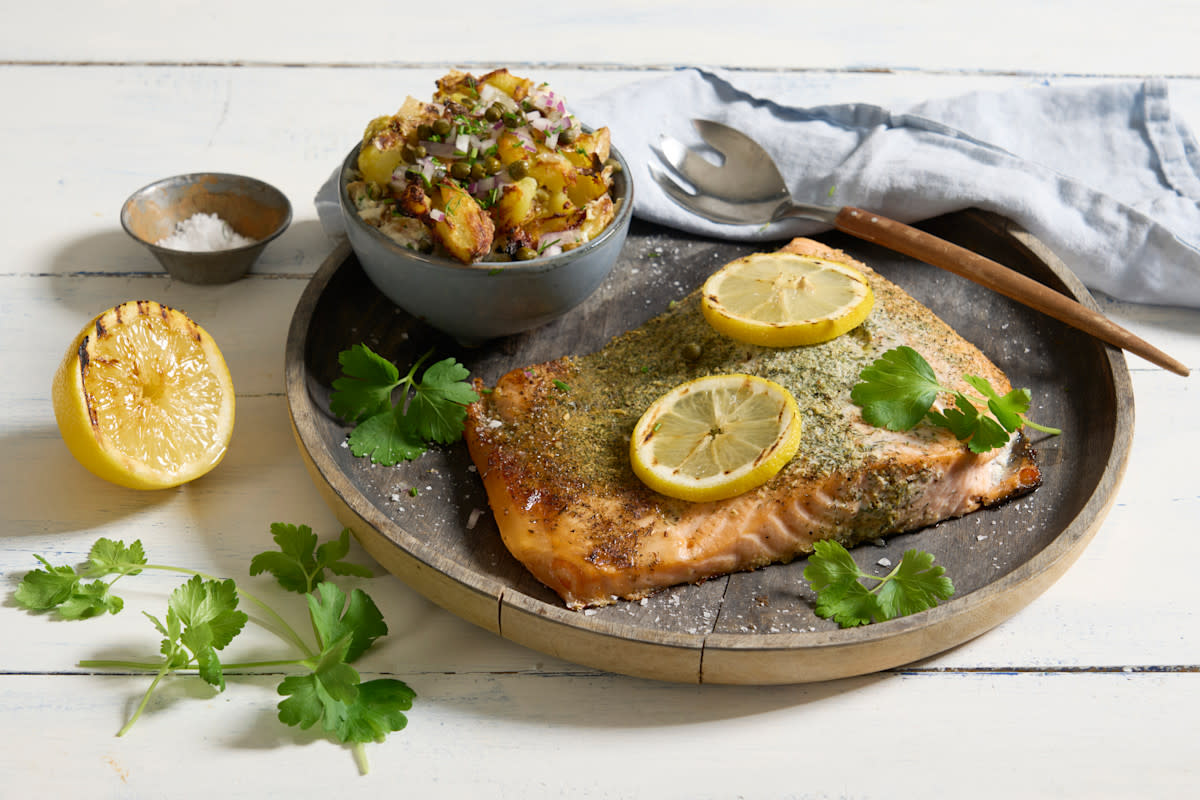 Grillet laks med urter og crispy smashed potetsalat servert på rundt trefat med sitronskiver over og liten skål potetsalat ved siden av.