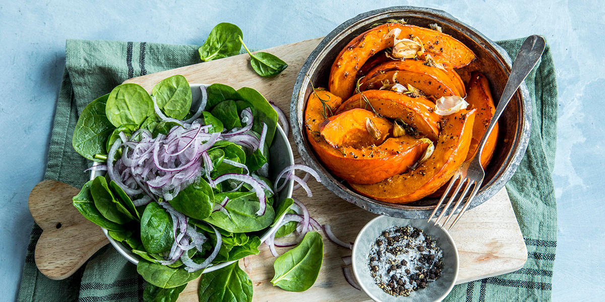 Til høyre en skål med store båter ovnsbakt gresskar med hele hvitløksfedd. Til venstre en skål med frisk spinat med tynne skiver rødløk. Liten skål med grovt salt og pepper.