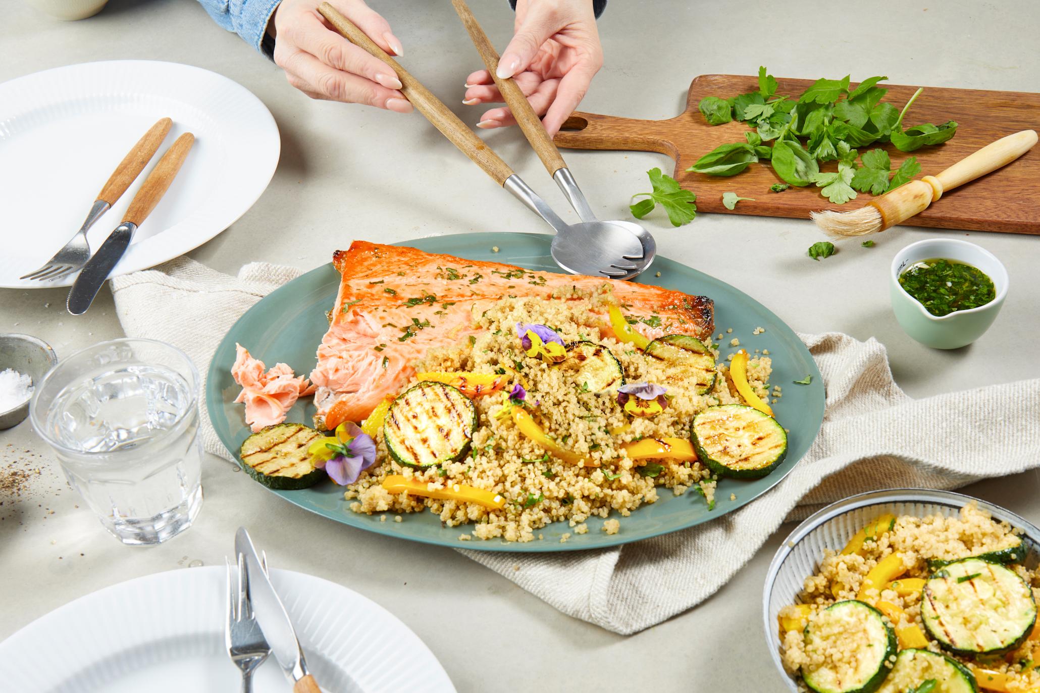 Grillet laks med quinoa, ferske urter og grillet squash servert på blågrønt fat på dekket middagsbord. To hender holder serveringsbestikk over fatet. Bolle med quinoa og grillet squash i forgrunn, og mørk trefjøl med grønne urter i bakgrunn.