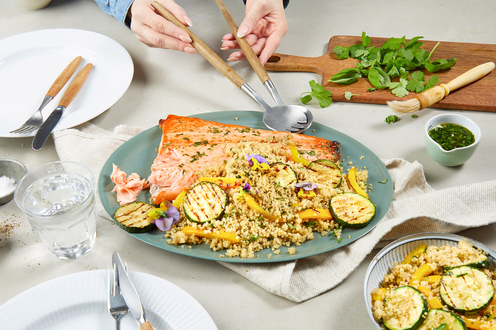 Grillet laks med quinoa, ferske urter og grillet squash servert på blågrønt fat på dekket middagsbord. To hender holder serveringsbestikk over fatet. Bolle med quinoa og grillet squash i forgrunn, og mørk trefjøl med grønne urter i bakgrunn.