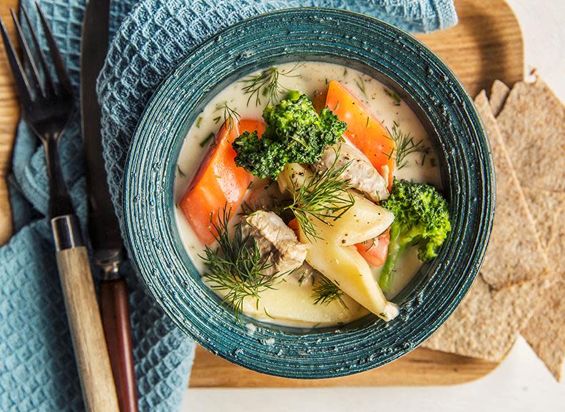 Lammegryte med rotgrønnsaker og dill i lys saus servert i mørk turkis bolle med flatbrød og bestikk ved siden av. Gryten er pyntet med kruspersille.