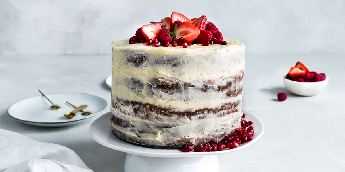 Saftig gulrotkake med kremostglasur lagt i fire lag, og pyntet med jordbær og granateplefrø, servert på hvitt stettfat.