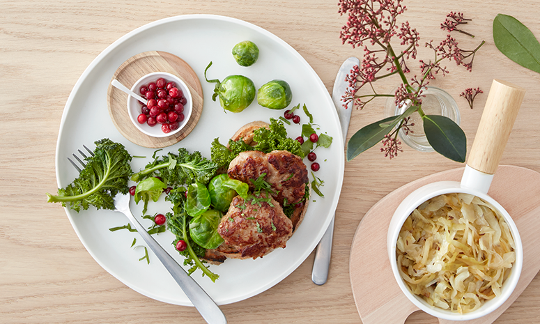 Medisterkaker med grønnkålsalat servert på et stykke brød med en liten skål ferske tyttebær ved siden av. Brødskiven står på hvit tallerken på lyst trebord.