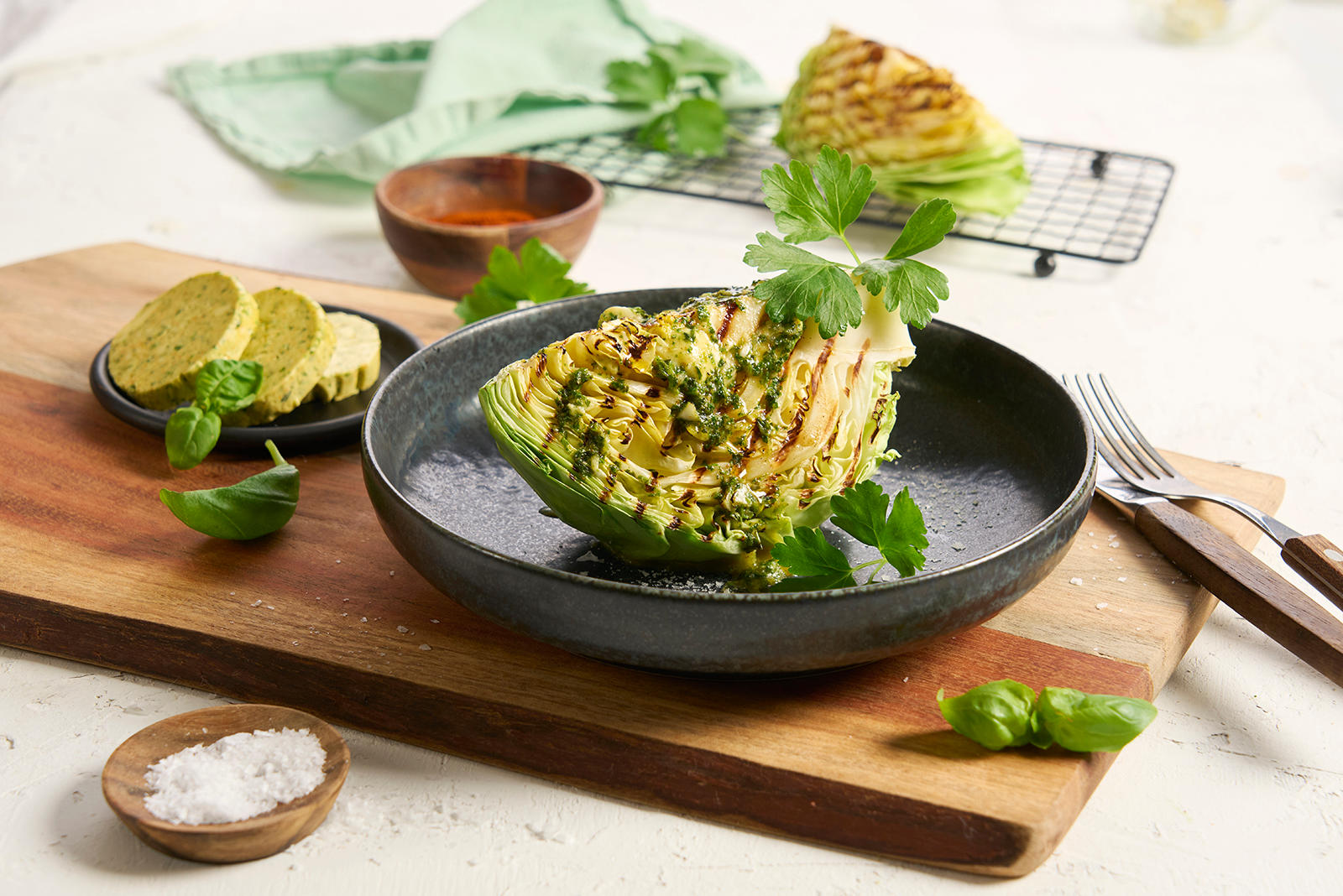 Grillet nykål med hjemmelaget kryddersmør servert med friske urter på mørk tallerken på trefjøl med skiver av kryddersmør ved siden av. Grillet båt med nykål i bakgrunnen.