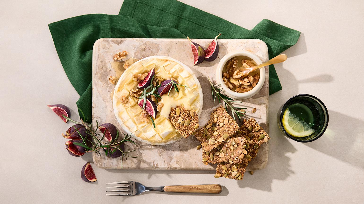 Smeltet brie på beige marmorfjøl, omringet av ferske fiken, rosmarin, valnøtter i honning og kjeks med frø og kjerner.