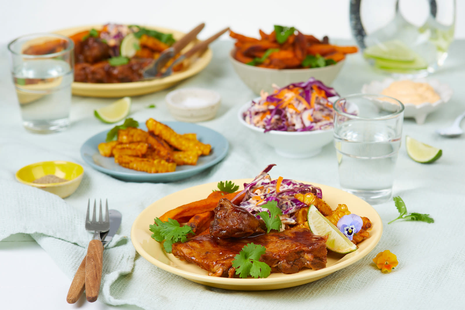 Baby Back Ribs med søtpotetfries, coleslaw og cornribs på en gul tallerken med diverse fat i bakgrunnen med mat på.