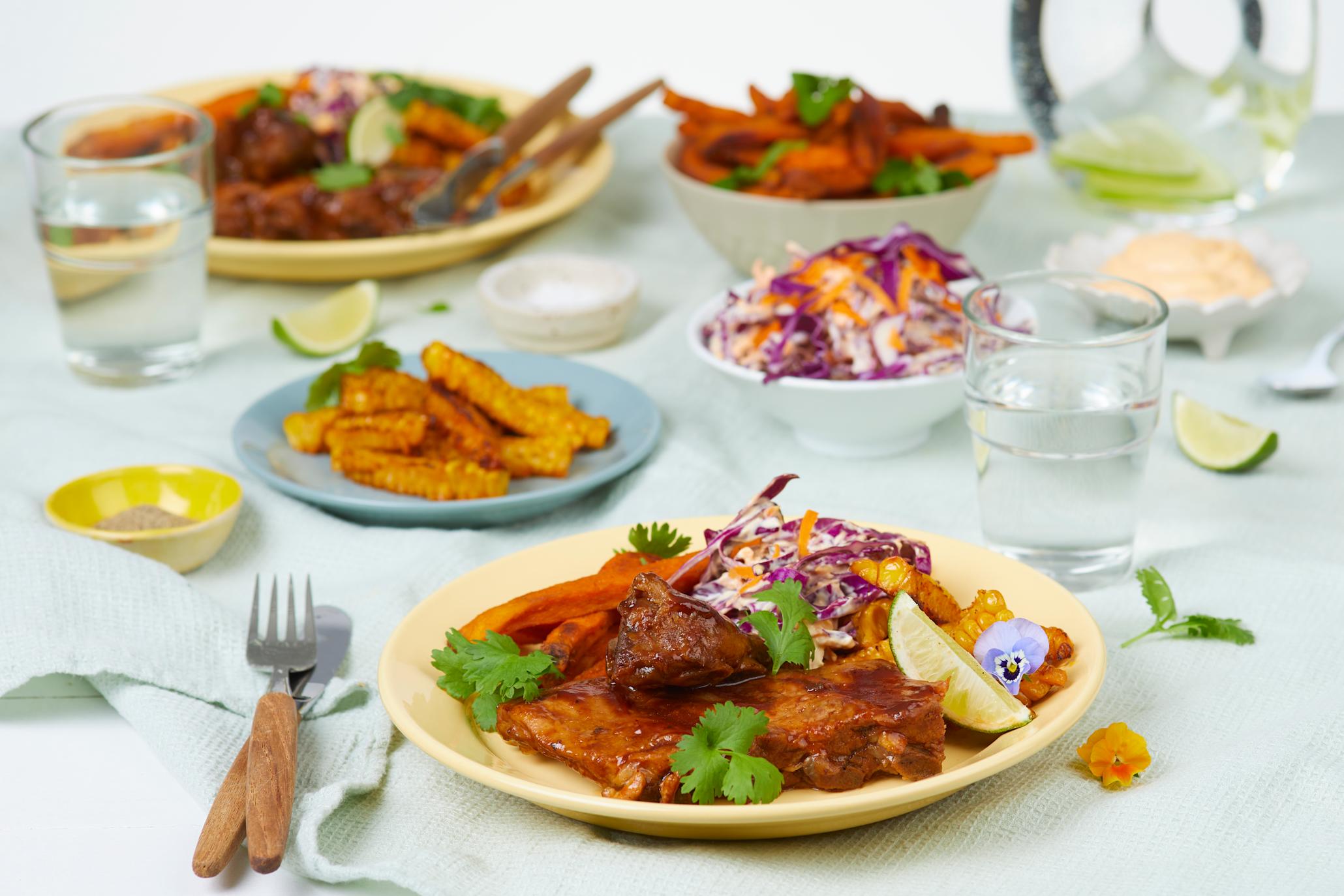 Baby Back Ribs med søtpotetfries, coleslaw og cornribs på en gul tallerken med diverse fat i bakgrunnen med mat på.