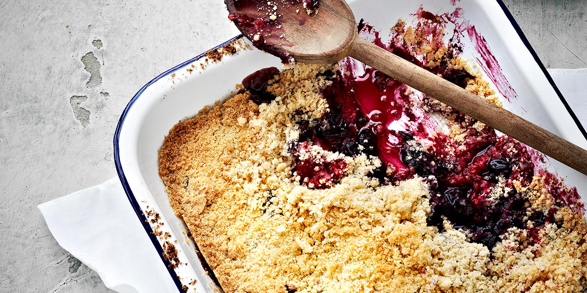 Hvit ildfast form med smuldrepai med bær. Det er tatt en stor skje av desserten, og en tresleiv ligger på toppen av formen.