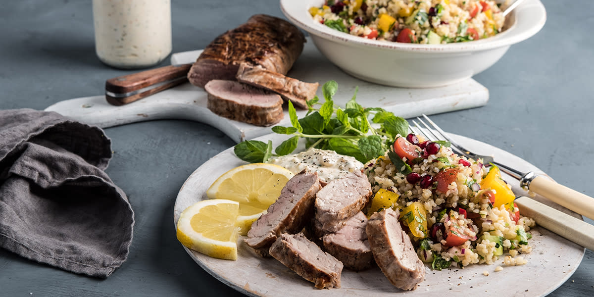 Helgrillet indrefilet av svin med fargerik bulgursalat og fetadressing, servert på hvit tallerken med sitronskiver og fersk basilikum. I bakgrunnen er det en skål med mer bulgursalat og en krukke med fetadressing, samt en halv grillet indrefilet.