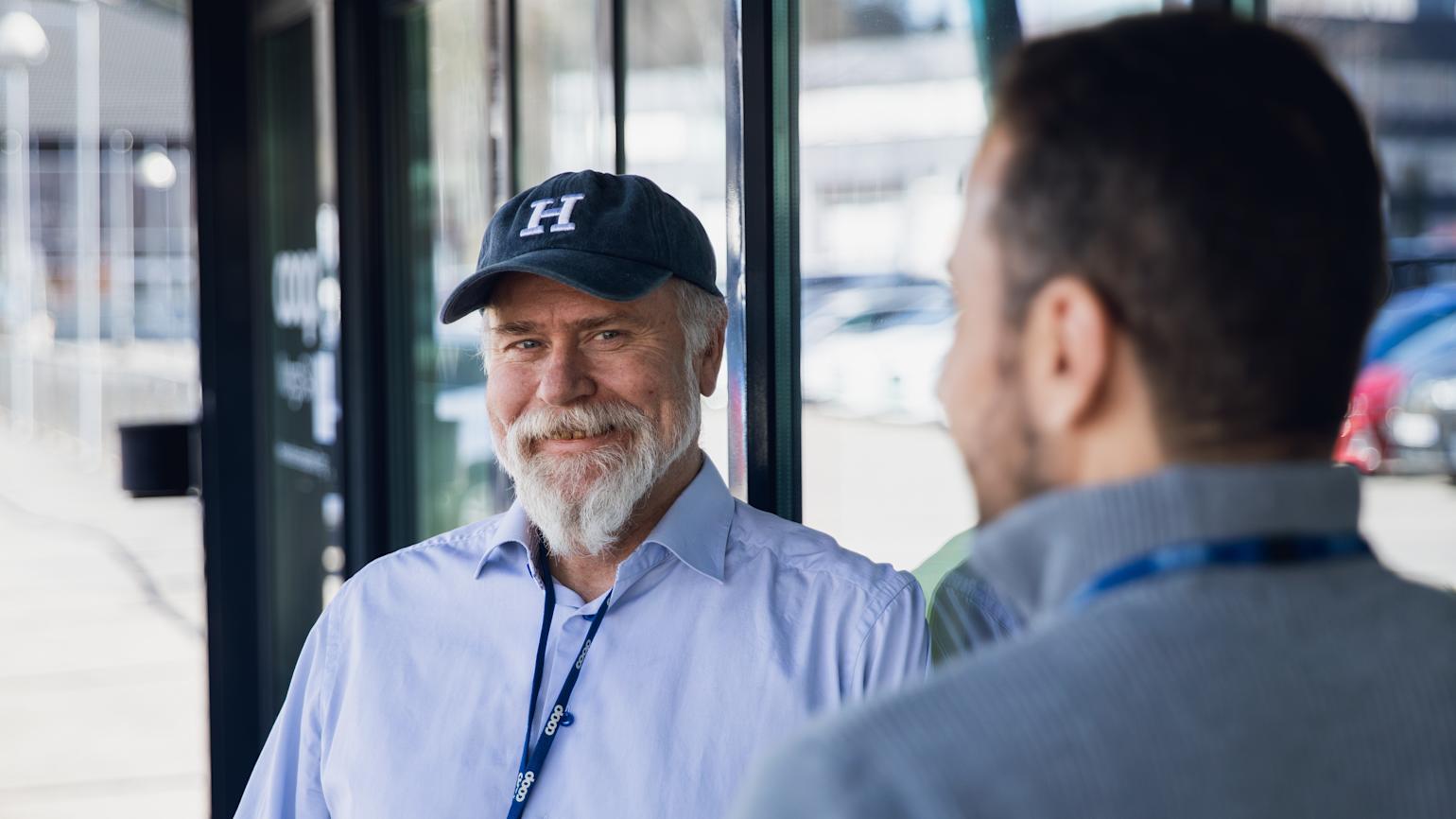 To menn lener på en glassvegg utenfor et kontor. Mannen til venstre har en Caps på seg og en nøkkelkort hengende rundt halsen, den andre mannen har ryggen til kamera