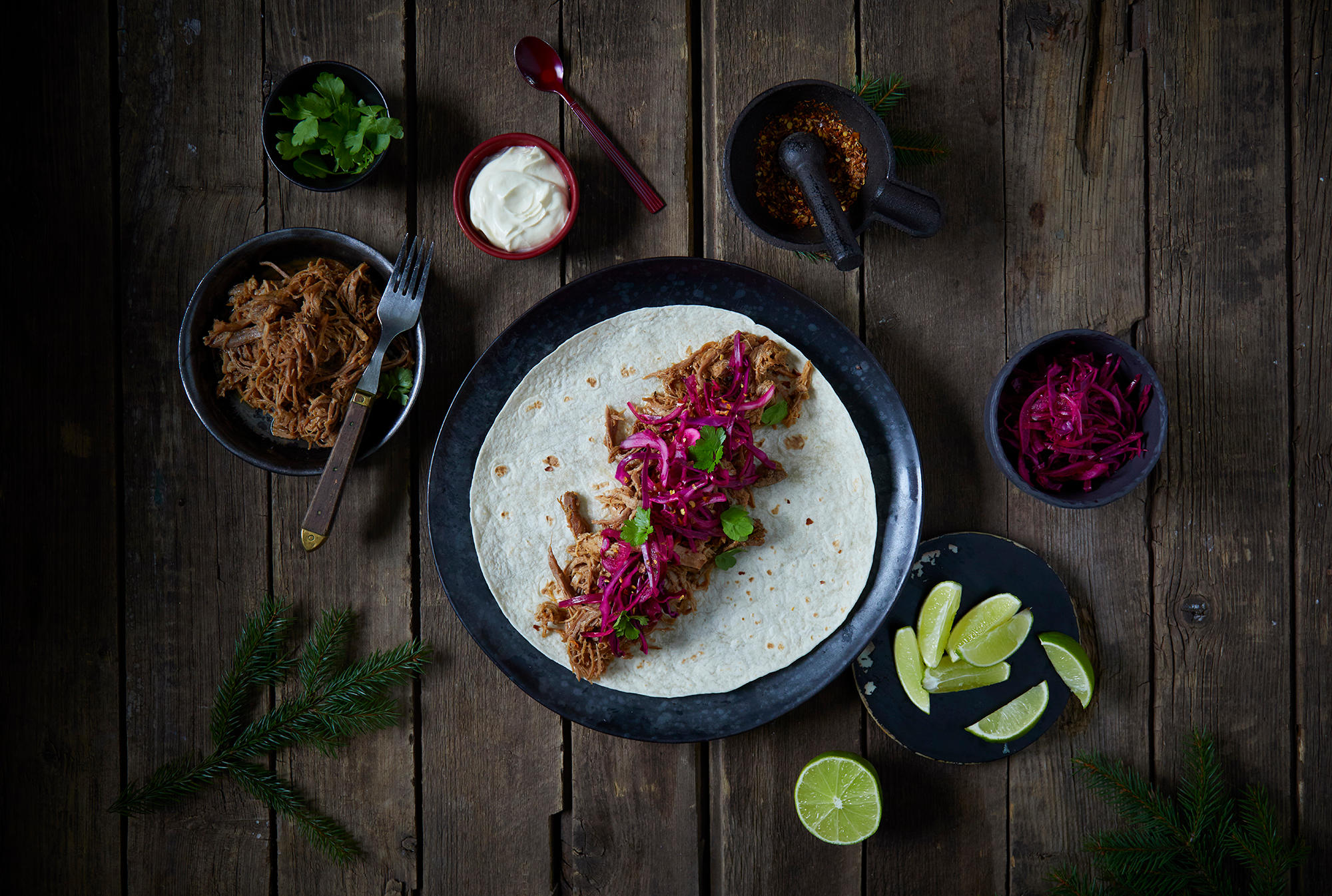 Taco med pulled ribbe og rødkålsalat servert på tortillalefse på svart tallerken. Tallerkenen står på rustikk trebord, med mørke skåler med tilbehør rundt. Granbar på siden.