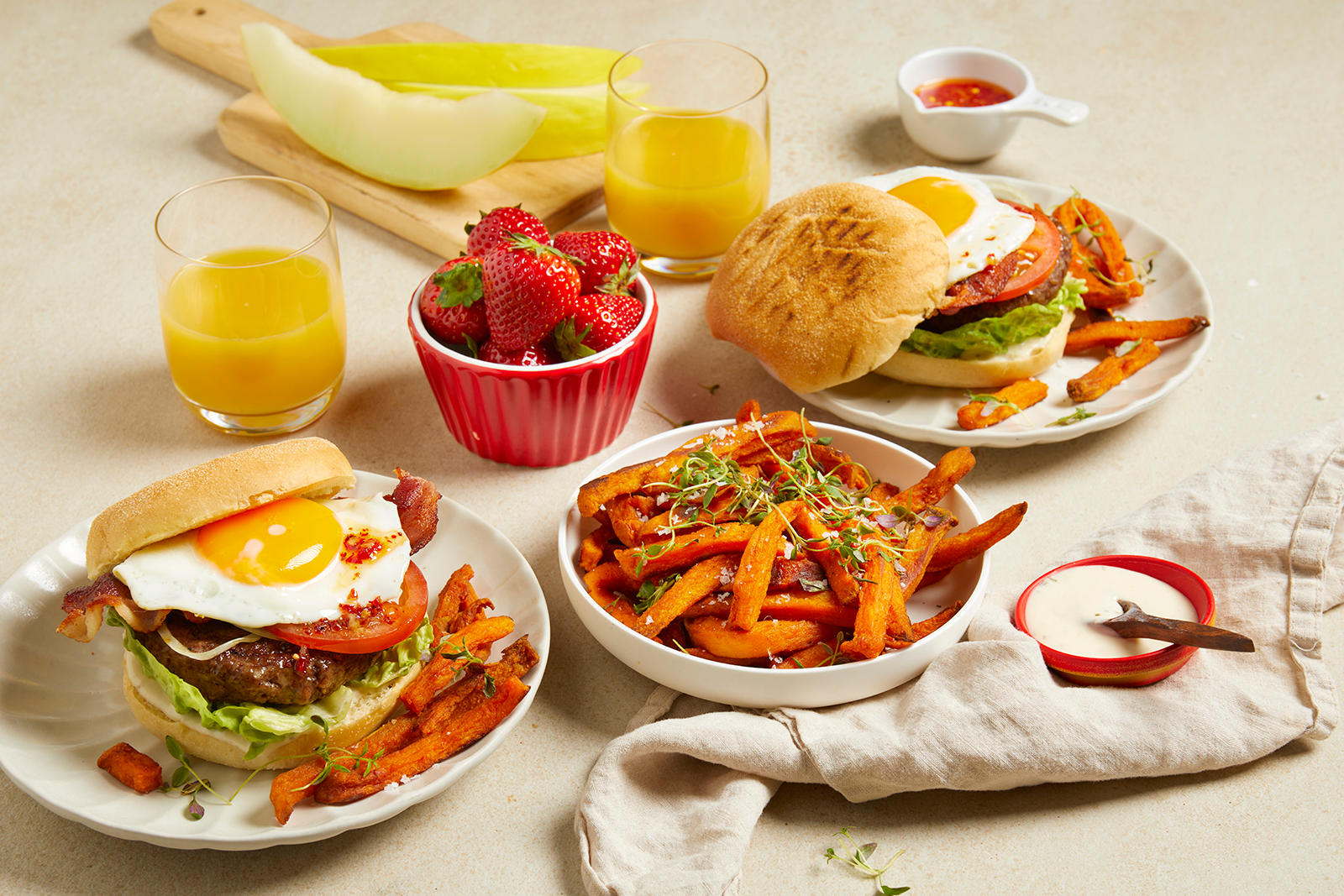 Brunch burger med speilegg og kjapp chiliolje, tomat, bacon og salat, servert på hvite asjetter på beige bakgrunn. Skåler med søtpotetfries, jordbær og chiliolje ved siden av. Glass med appelsinjuice, og skiver av honningmelon i bakgrunnen.