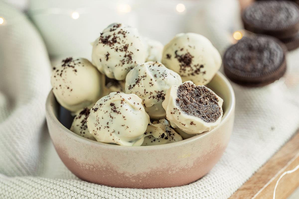 Mørke marsipankuler med Oreo trukket i hvit sjokolade, med et dryss Oreosmuler på toppen. Servert i lys skål, på lys stoffbakgrunn. Oreos og julelys i bakgrunnen.