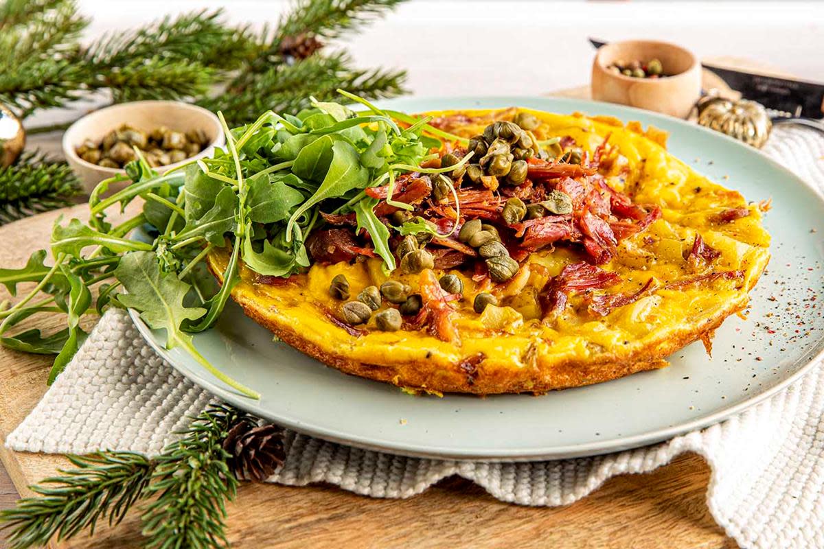 Gylden omelett med potetbiter og revet pinnekjøtt, toppet med kapers og ruccola. Servert på lyst fat. Rustikk trebakgrunn og granbar.