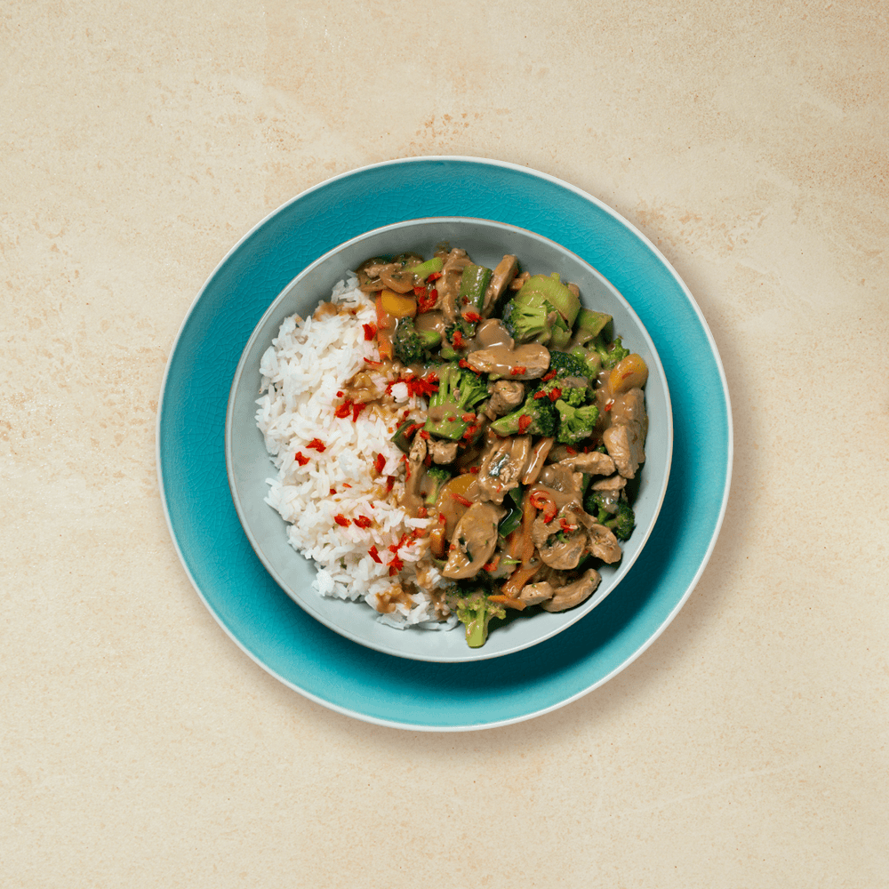 Asiatisk hverdagsgryte med svin, wokgrønnsaker og peppersaus, servert i lys blå skål med ris og finhakket rød chili. Skålen står på turkis tallerken, på beige bakgrunn.