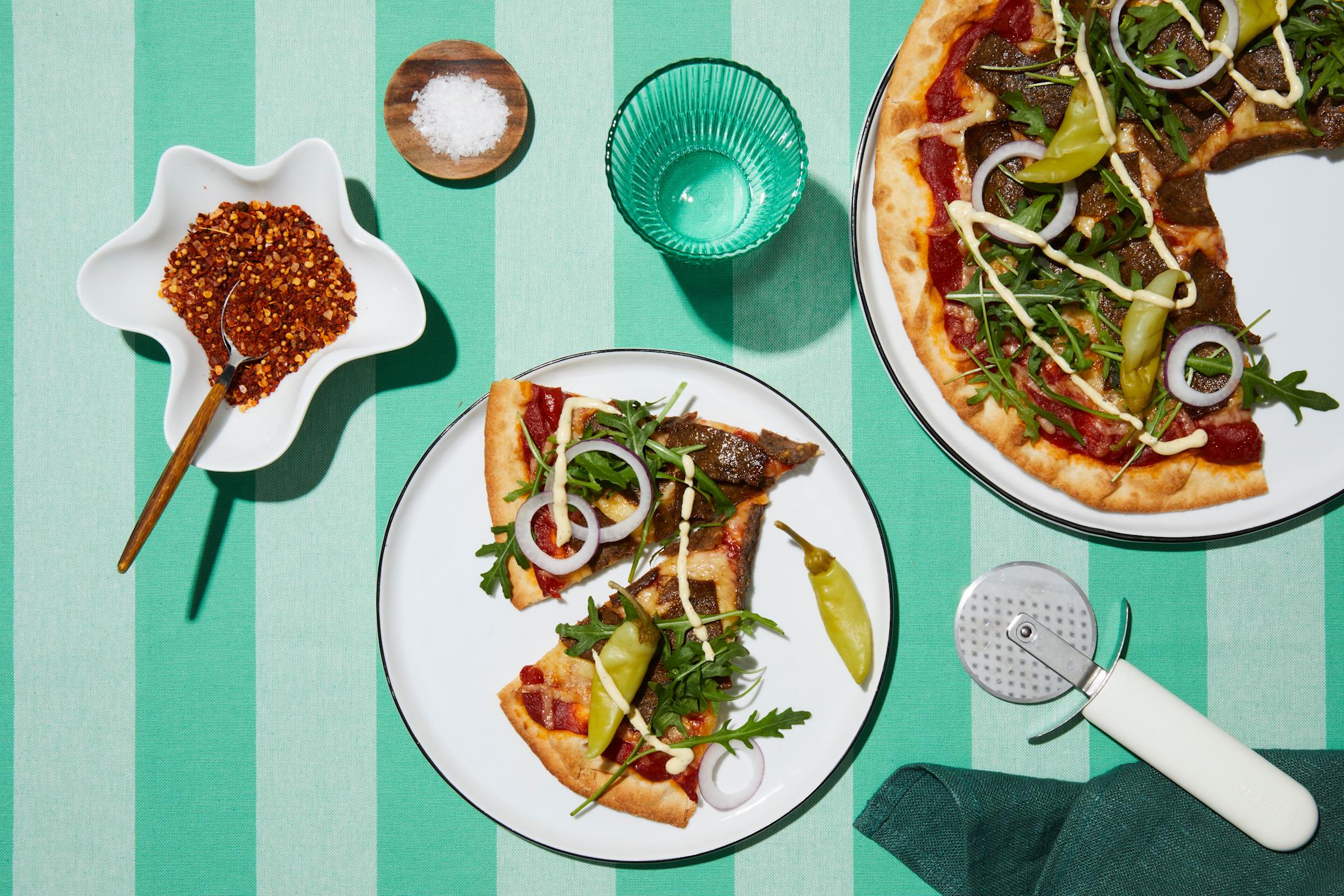 Kebabpizza med ruccolasalat, rødløk, kebabdressing og syltet chili servert på hvit tallerken på turkis stripet duk. Små skåler med krydder på siden.