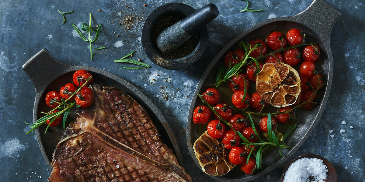 Ovnsbakte piccolotomater og hel hvitløk delt i to, toppet med fersk rosmarin og servert i svart ildfast form. Ved siden av er en ildfast form med grillet t-beinstek.