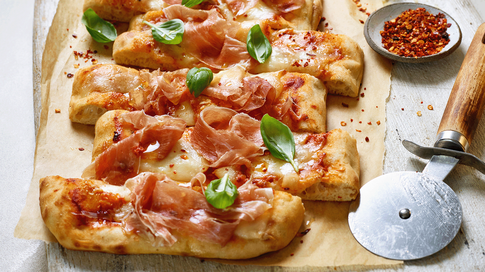 Pizza prosciutto toppet med basilikum er delt i skiver,  og ligger på bakepapir med et pizzahjul og en liten skål med chilliflak ved siden av.