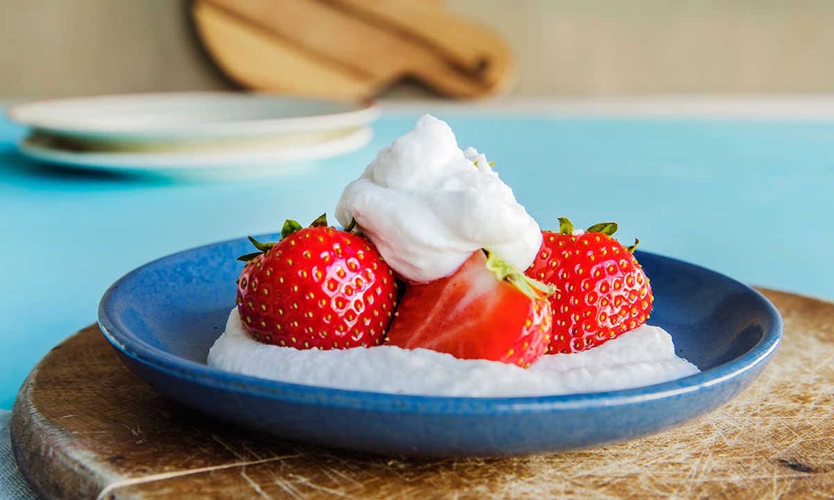 Vegansk pisket krem av kokosmelk servert i mørkeblå skål med hele og delte friske jordbær. Skålen står på rund trefjøl på blått bord.